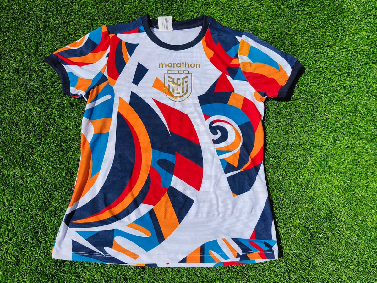 A short-sleeve athletic jersey with a bold abstract geometric pattern in white, navy, orange, and blue, laid flat on green grass. The jersey features the 'marathon' brand name and a crest on the chest.
