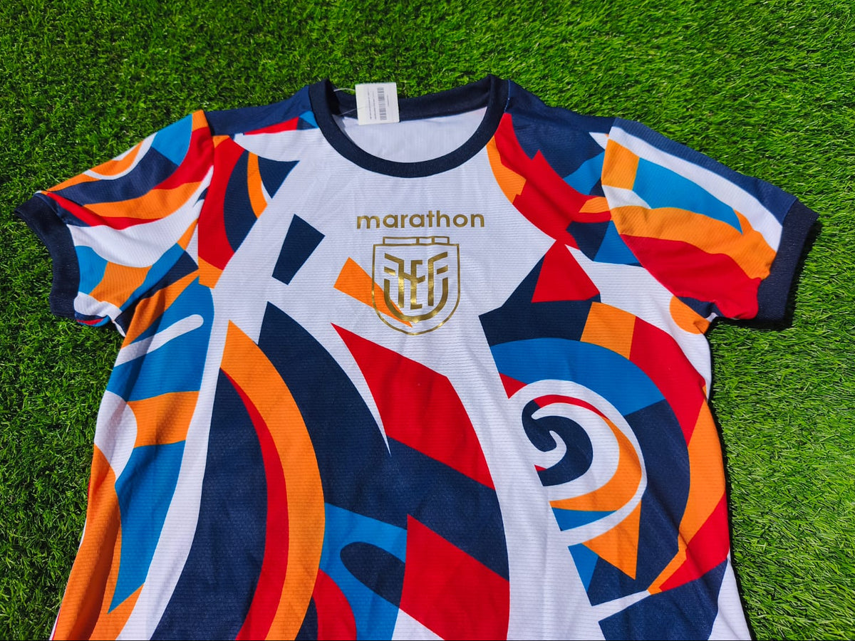 A short-sleeve athletic jersey with a bold abstract geometric pattern in navy, white, red, and orange, laid flat on green grass. The jersey features the word 'marathon' and a crest on the chest.